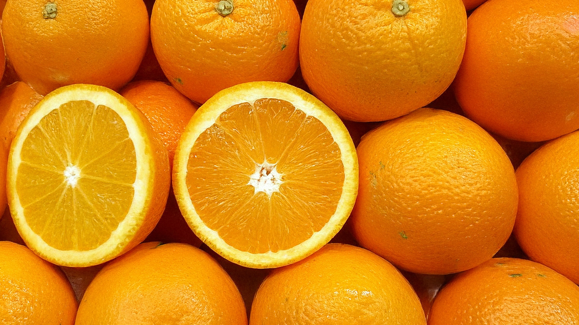 Großaufnahme von etwa 15 ganzen Orangen. Eine ist aufgeschnitten, sodass das Fruchtfleisch der zwei Hälften zu sehen ist. 