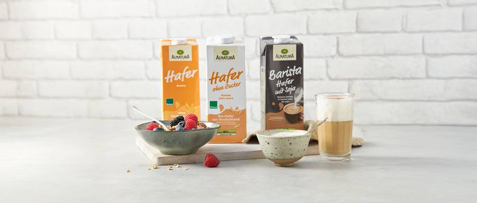 Das Bild zeigt drei Kartons der Alnatura Hafergetränke: Haferdrink, Haferdrink ohne Zucker und Barista Haferdrink mit Soja. Daneben stehen eine Schale mit Müsli und Beeren sowie zwei Getränkegläser, eines mit einem mit Haferdrink zubereiteten Cappuccino oder Latte Macchiato. Der Hintergrund ist eine weiße Backsteinwand. Das gesamte Arrangement wirkt hell, freundlich und appetitanregend und soll die Vielseitigkeit der Alnatura Hafergetränke hervorheben.