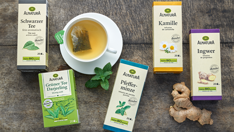 Es liegen fünf Alnatura Tee-Verpackungen von den Sorten Schwarzer Tee, Kamille, Ingwer, Pfefferminze und Grüner Tee auf einem Holzuntergrund mit einer Tasse Tee mit Beutel und Minze und eine Ingwerknolle 