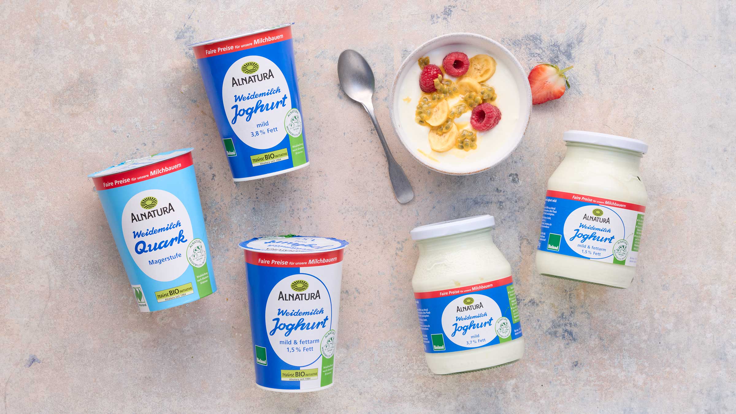 Das Bild zeigt verschiedene Produkte der Marke Alnatura: Joghurt (mild, 3,8% Fett; mild & fettarm, 1,5% Fett) und Magerquark.  Die Produkte sind auf einem hellen Untergrund arrangiert, neben einer Schale Joghurt mit Früchten (Himbeeren, Banane, Passionsfrucht) und einem Löffel.  Das Bild soll die Produkte präsentieren.