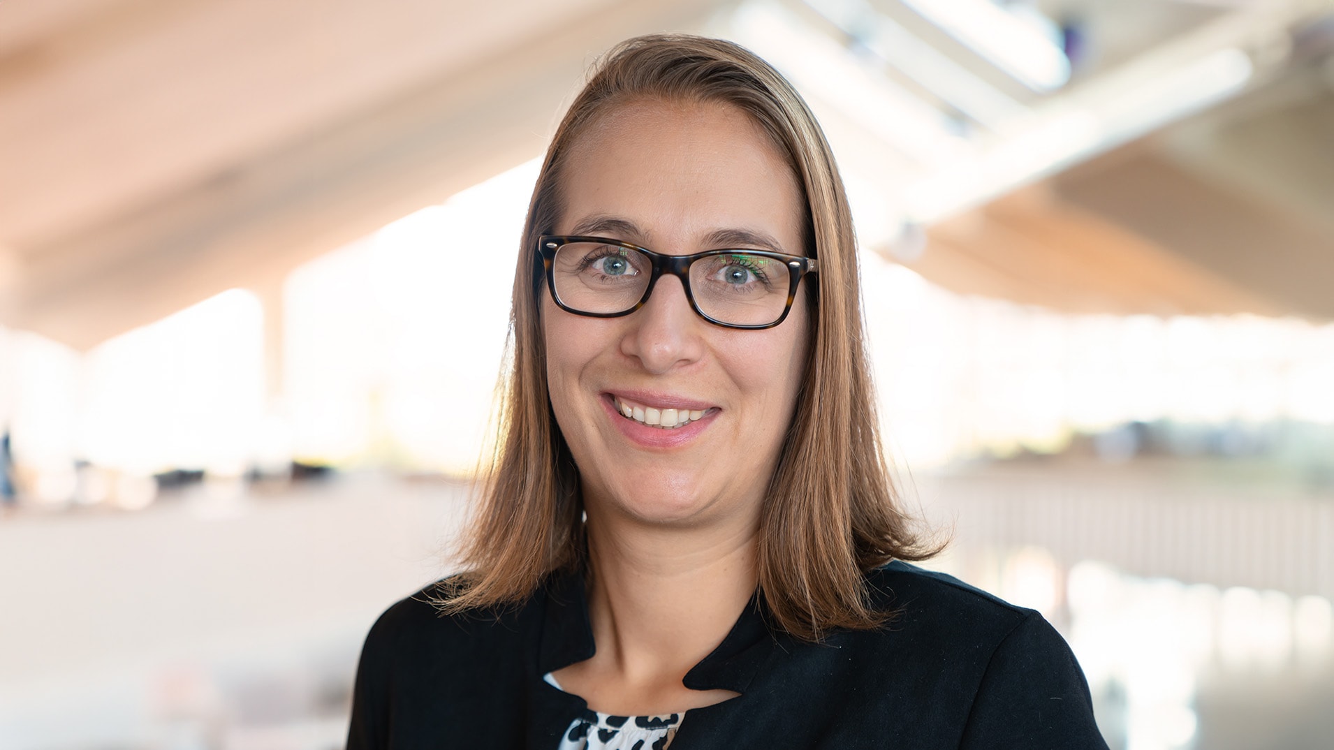 Eine Frau mit Brille und halblangen Haaren lächelt in die Kamera. Die Frau ist im Business-Style gekleidet. Man sieht sie ab den Schultern aufwärts. Im Hintergrund ist verschwommen das innere eines Gebäudes zu sehen.